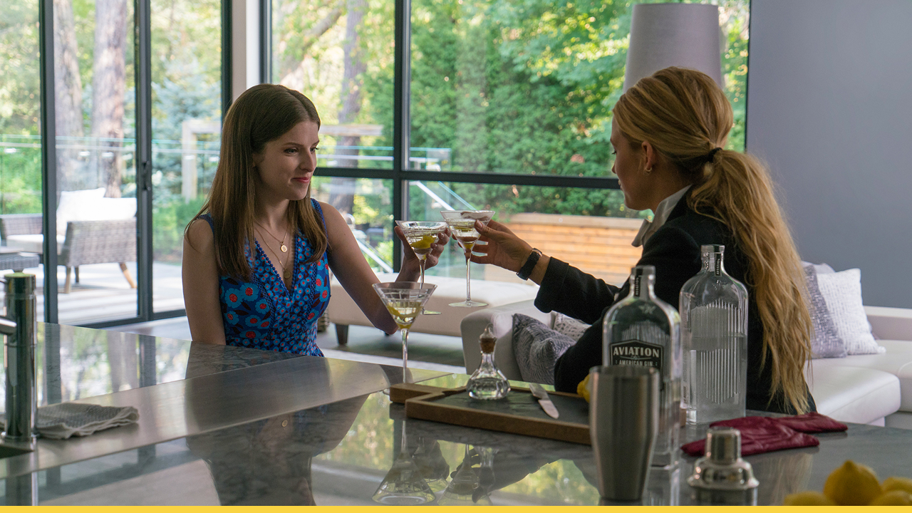 Two characters from A Simple Favor clinking martini glasses in a modern kitchen