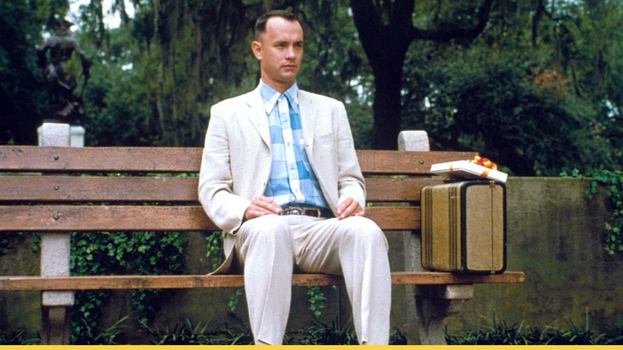 Forrest Gump in a light suit sits on a park bench with a suitcase and a box of chocolates.
