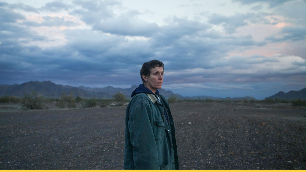 A woman stands alone in a vast desert landscape under a cloudy sky.