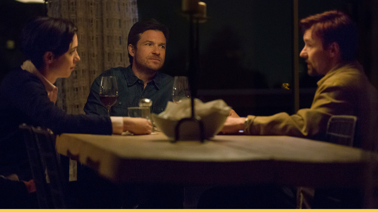 A dimly lit dinner scene from the movie The Gift, showing three characters—two men and a woman—seated around a table with wine glasses and a candle between them, creating a tense and suspenseful atmosphere.
